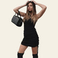 Woman wearing a black ruched drawstring mini dress, standing and holding a handbag