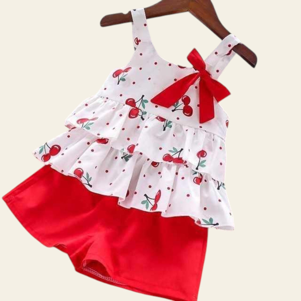 Children's outfit with white top and red shorts featuring cherry pattern on a wooden surface.