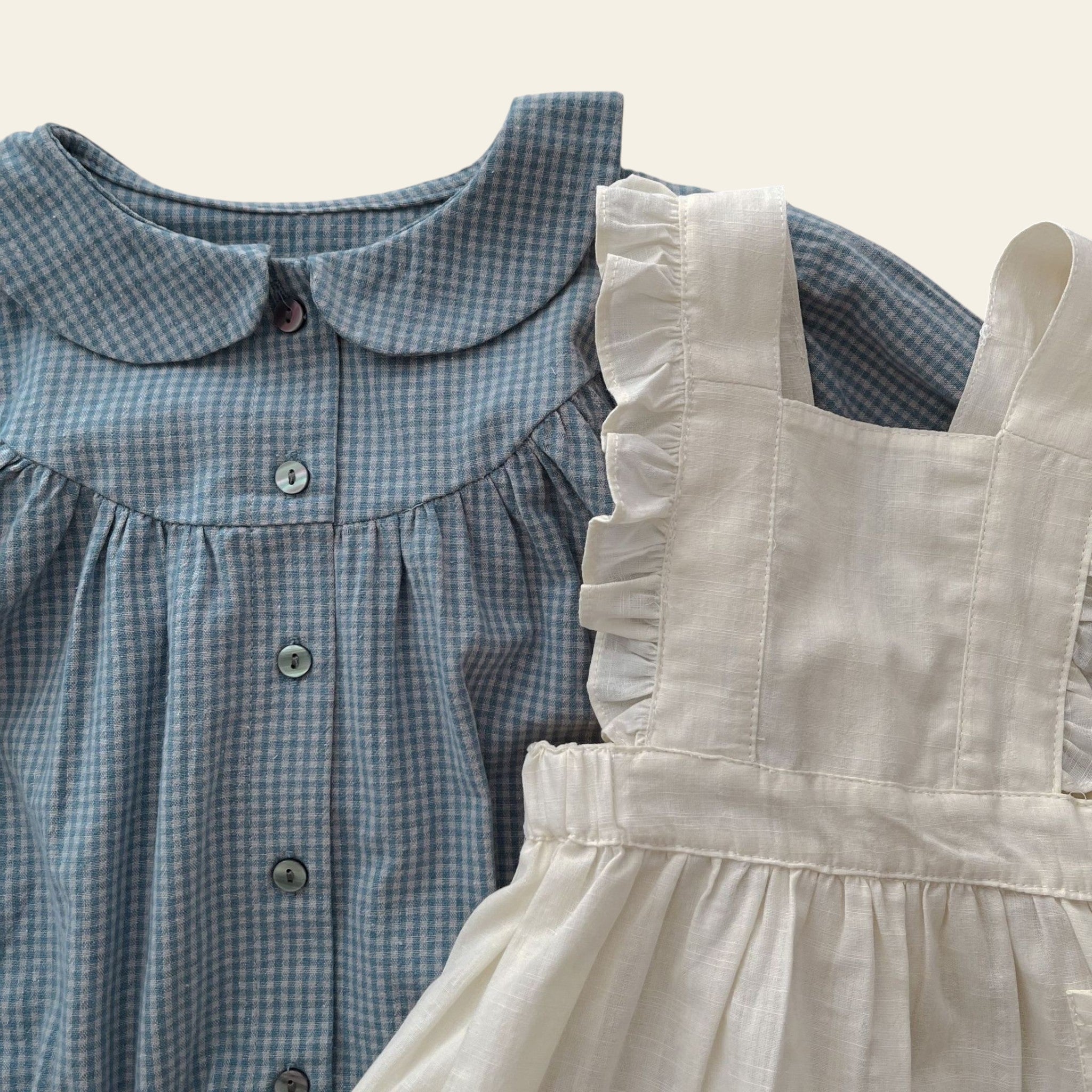 Two children's dresses, one blue checkered and one beige, on a wooden surface with an open book.