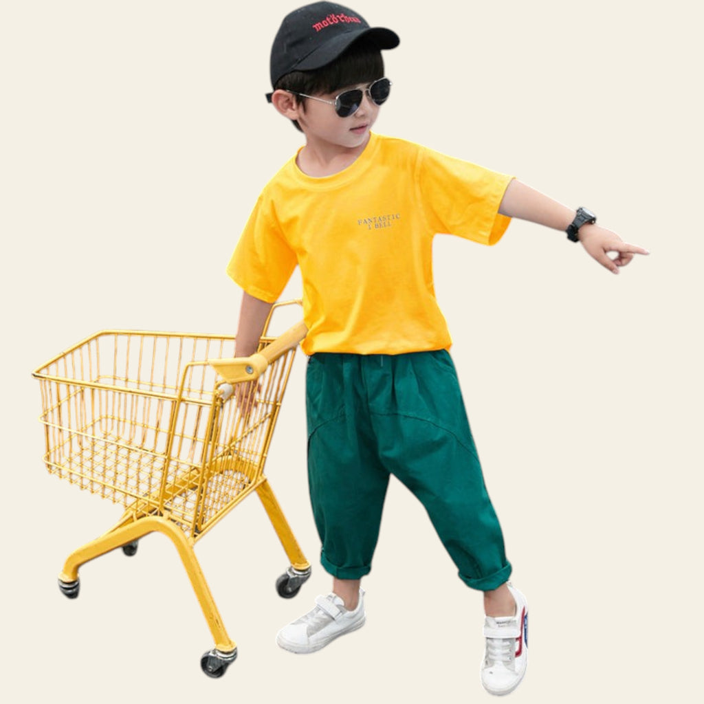 Child in yellow shirt and green pants standing next to a small yellow shopping cart on a dark surface.