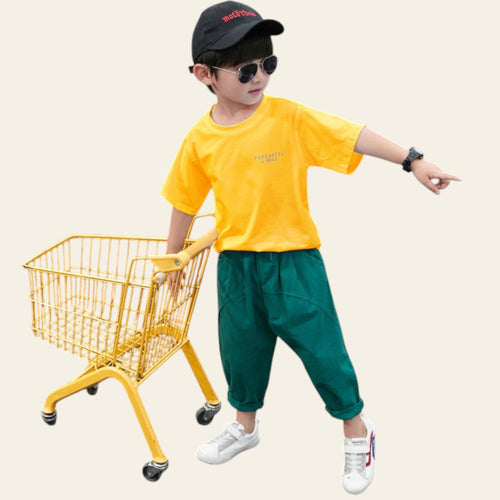 Child in yellow shirt and green pants standing next to a small yellow shopping cart on a dark surface.