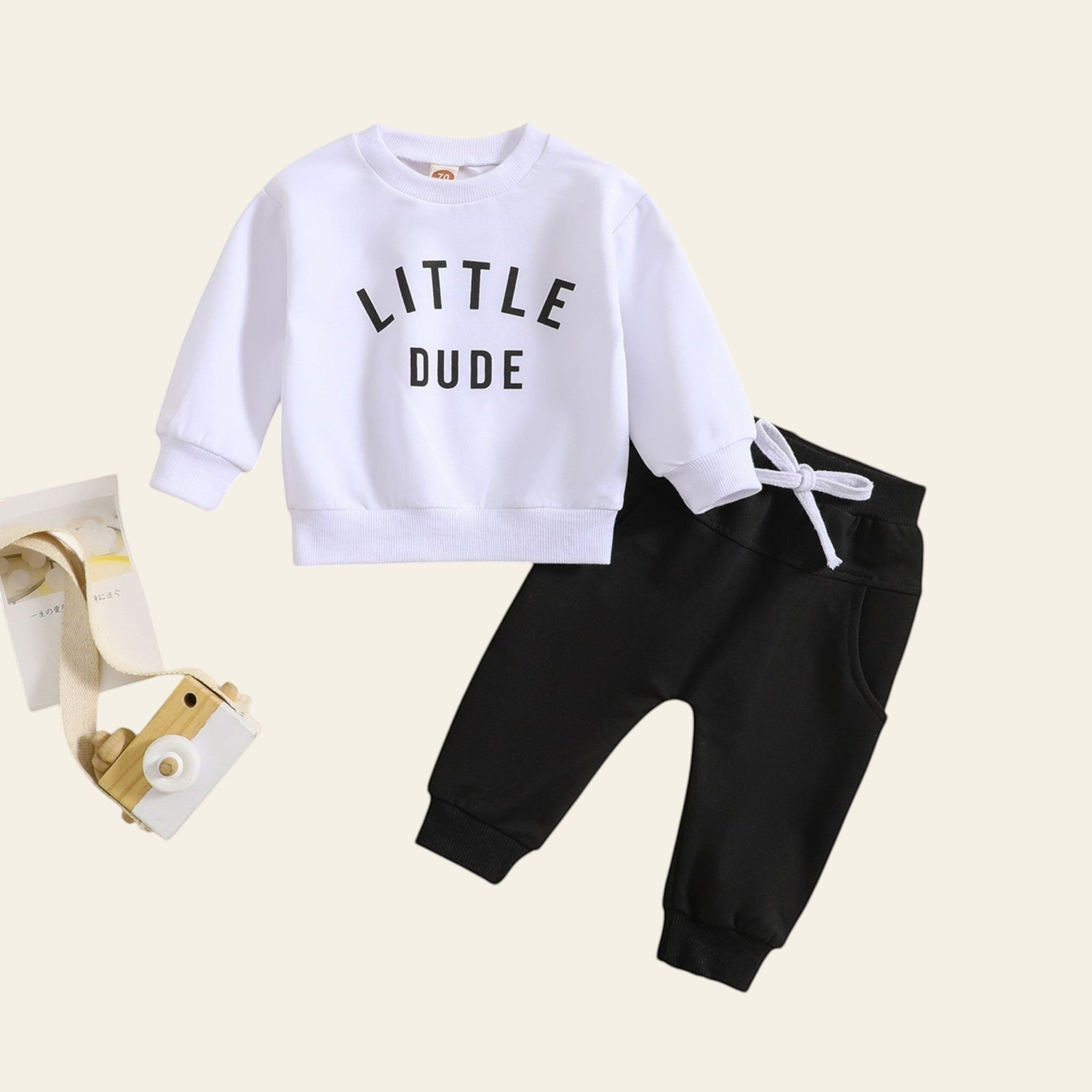 Children's white sweatshirt with 'LITTLE DUDE' text and black pants on a beige background.