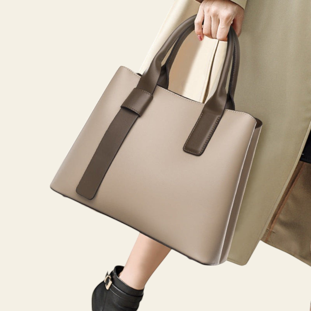 Beige handbag with brown handles held by a person wearing a beige coat and black boots.