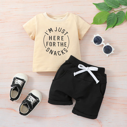 Children's outfit with t-shirt, shorts, shoes, and sunglasses on a light wooden surface.