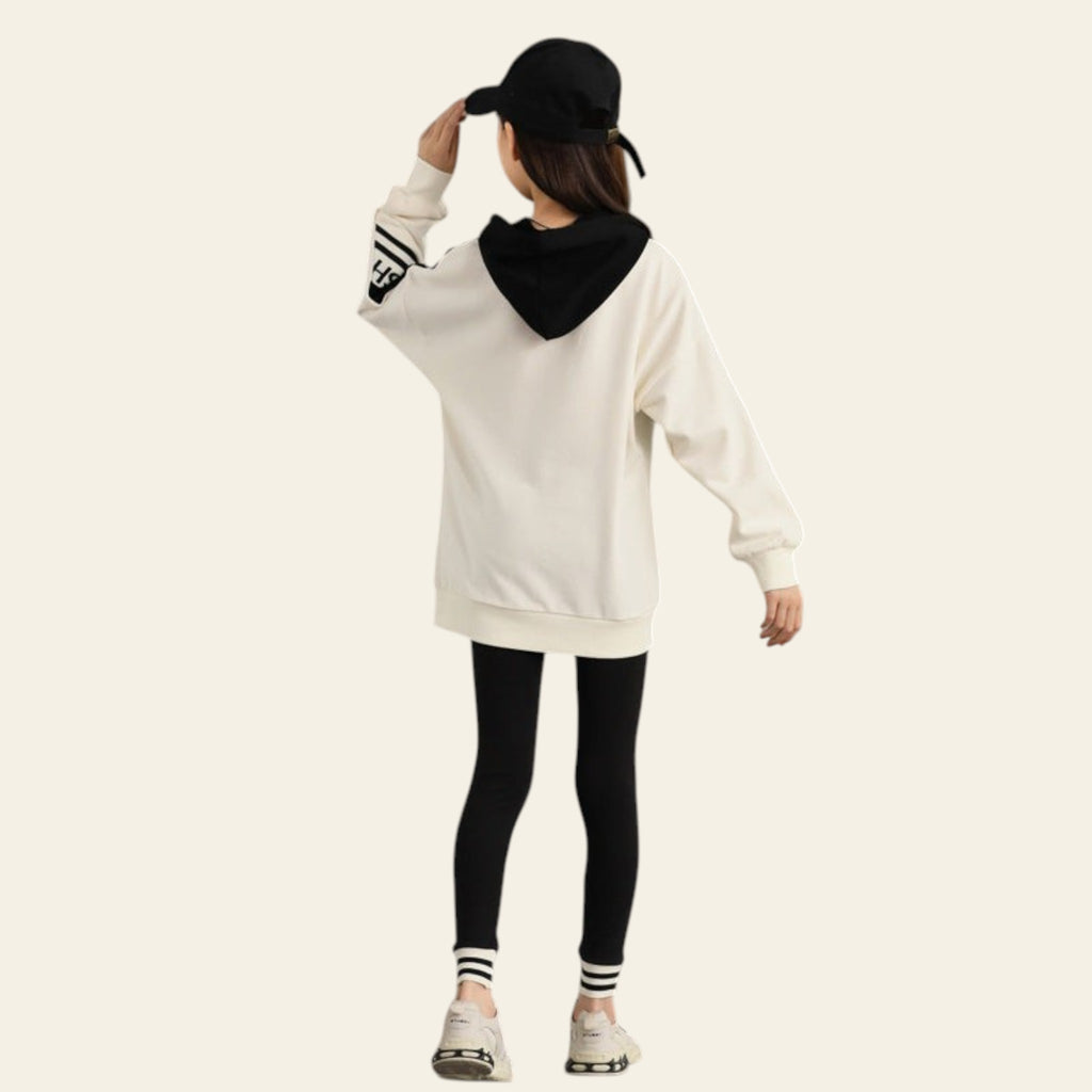 Person wearing a white hoodie with black hood and black leggings against a gray concrete wall.