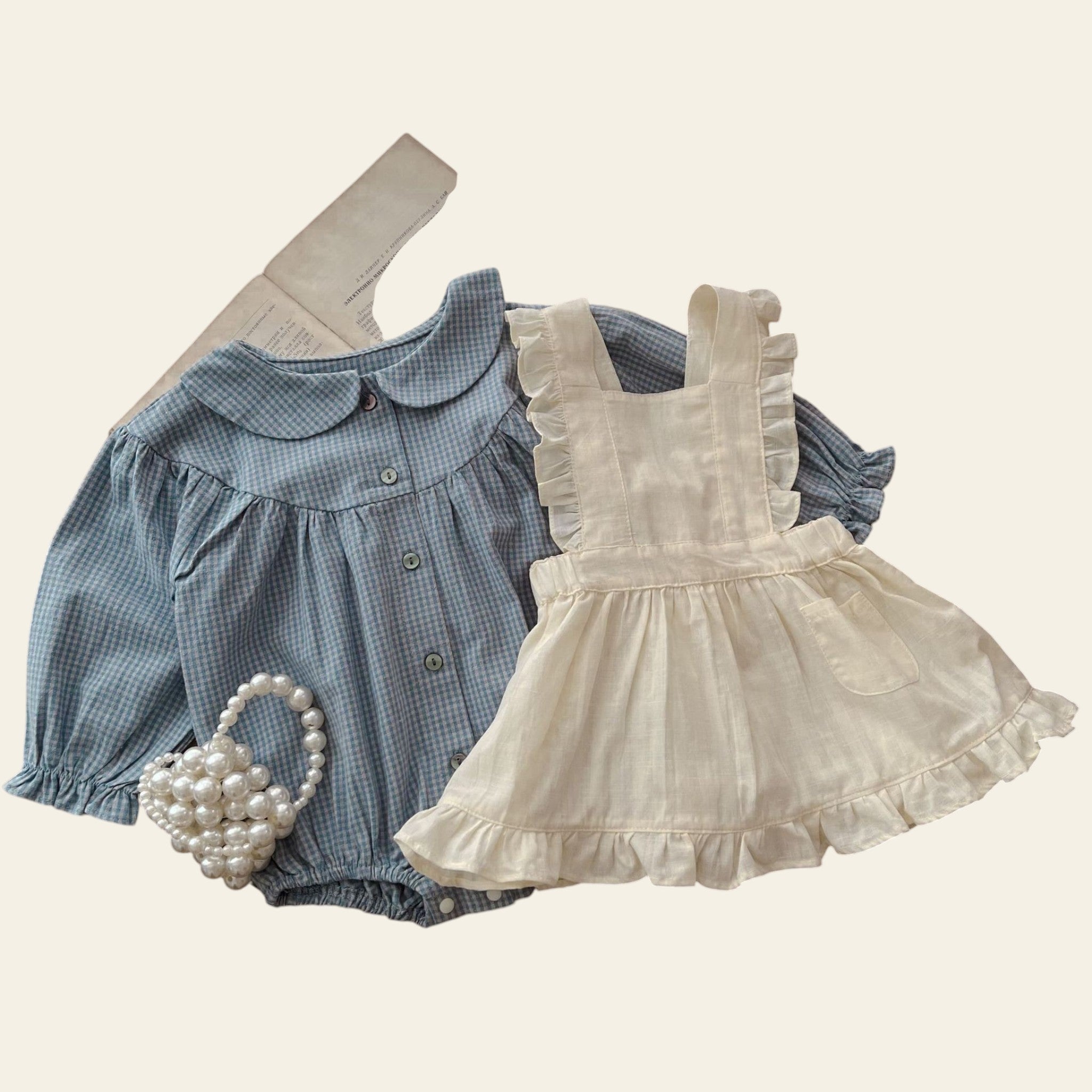 Children's blue checkered shirt and white dress on a wooden table with a pearl basket.