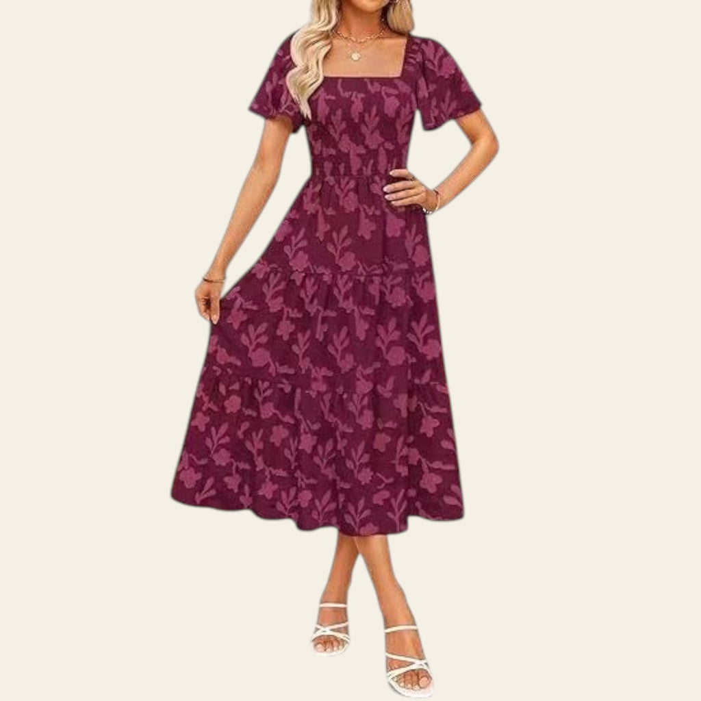 Woman wearing a purple floral dress standing outdoors with a white wall and pink geometric structure in the background.